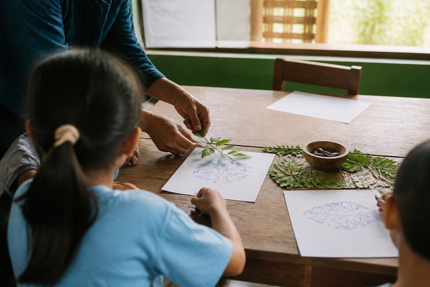 Tangan pendidik dan anak menata daun di atas meja untuk kegiatan eksplorasi bahan alam; wajah tidak terlihat.